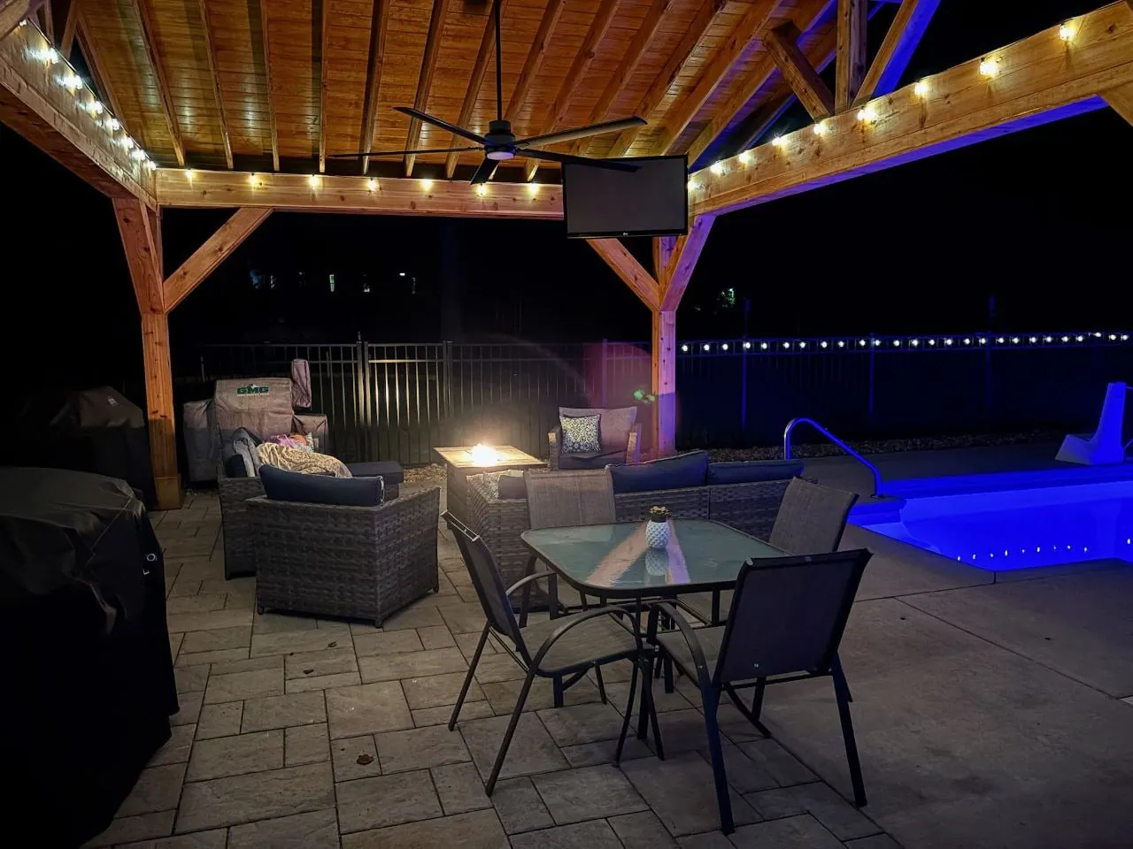 Covered timber pavilion at night with string lights, a ceiling fan and lounge seating beside a lit pool